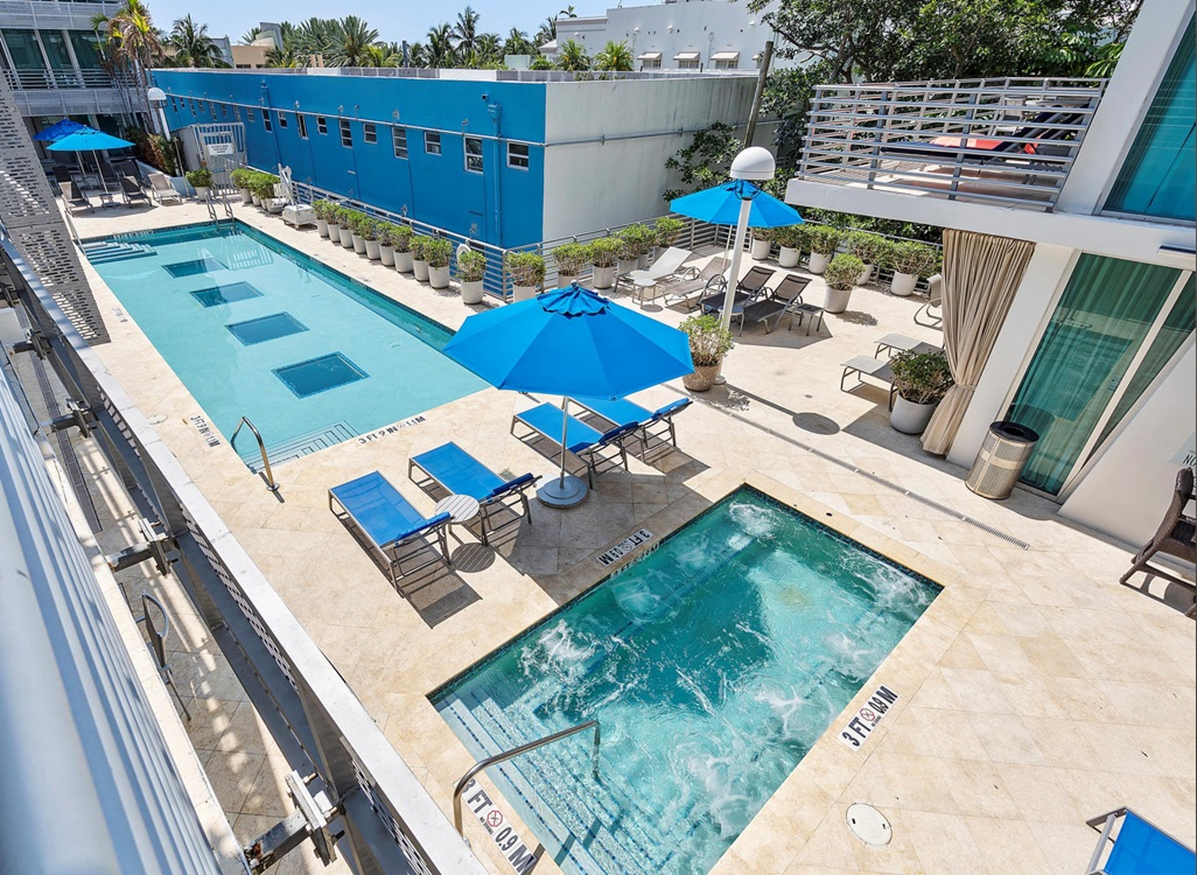 An aerial view of a hotel pool area with a rectangular pool, jacuzzi, lounge chairs, and umbrellas.